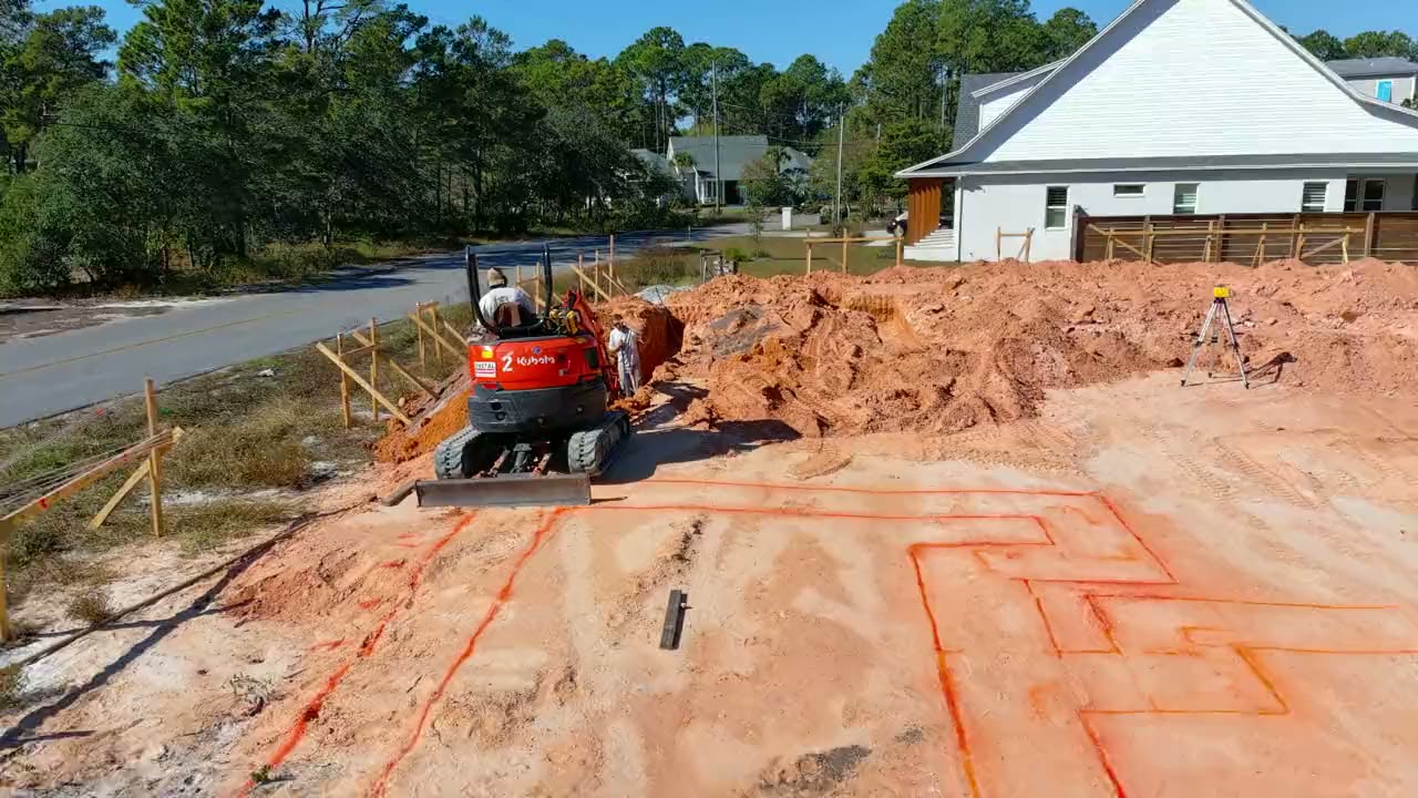 Aerial view of a cleared building lot with the foundation footprint marked out and a compact excavator working the ground.